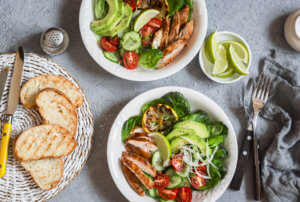 salad in a bowl with chicken and another plate with slices of toast. Wedges of lemon in a small dish on the right side. Fork on the right side of salad bowl.